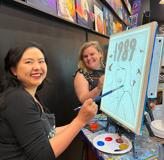 Two women smiling and painting a '1989 - Taylor Swift' canvas at Muse Paintbar. They are enjoying a creative painting session with brushes and vibrant paint colors at a fun, social art event.