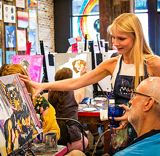 Instructor guiding a participant during a painting session at Muse Paintbar. Guests are enjoying a hands-on, creative experience in a vibrant studio filled with colorful artwork.