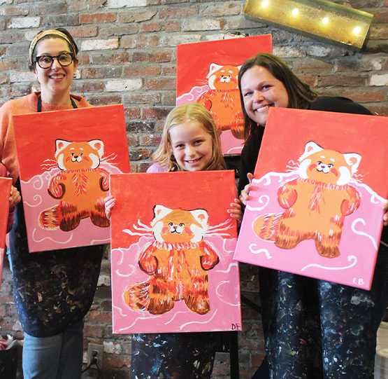 Family and friends proudly holding their red panda-themed paintings at Muse Paintbar. The group smiles together after completing a fun and creative art session at the vibrant studio.